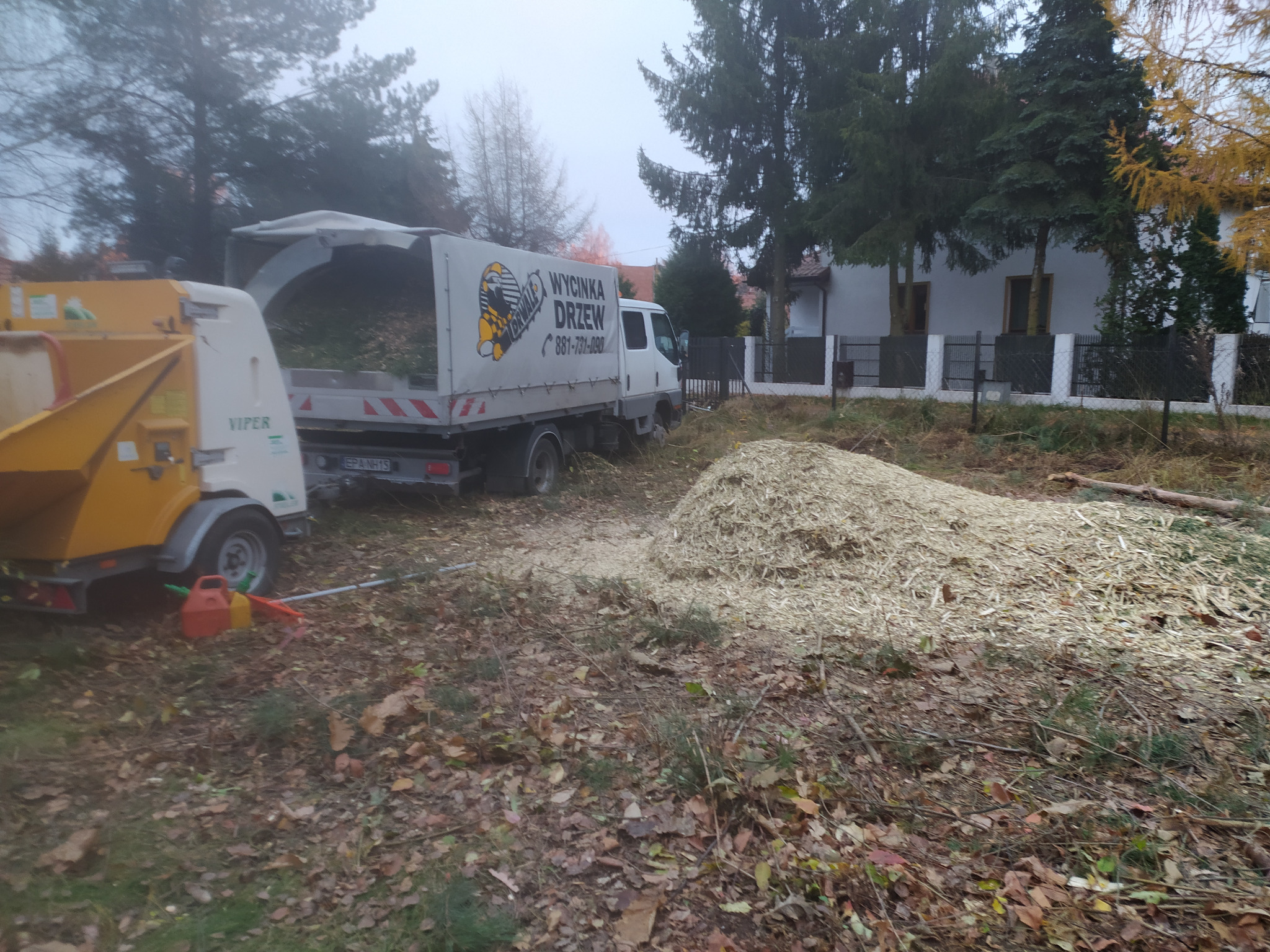 Ciężarówka z logo firmy zajmującej się obróbką drewna stoi obok żółtej rębarki, a na ziemi leży duża pryzma zrębków.
