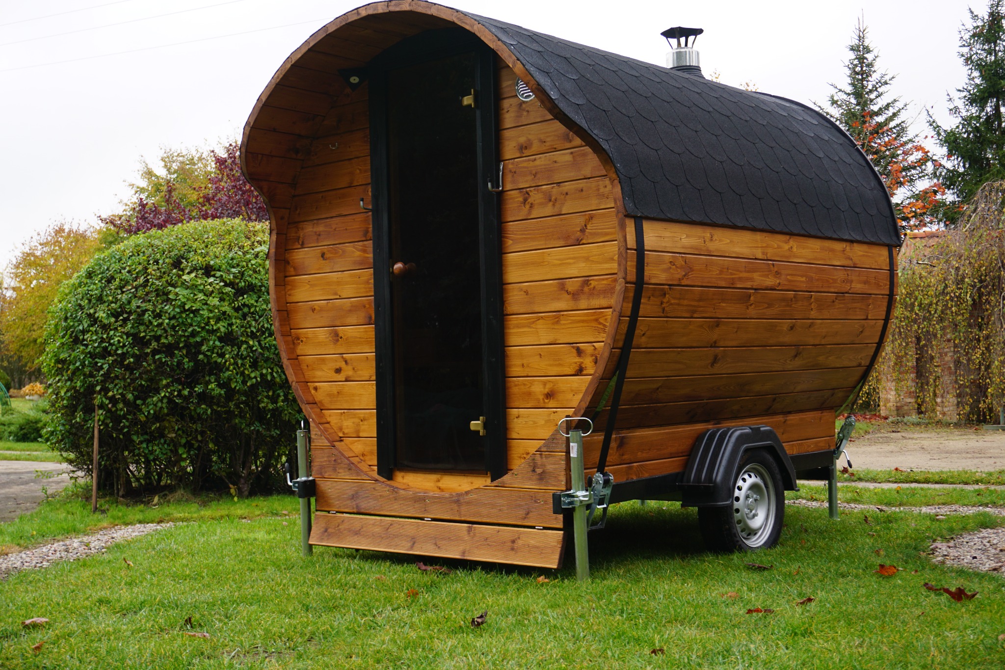 Mobilna, drewniana sauna w kształcie beczki na przyczepie w Starogardzie Gdańskim, z otwartymi drzwiami i czarnym dachem, ustawiona na trawie w ogrodzie.