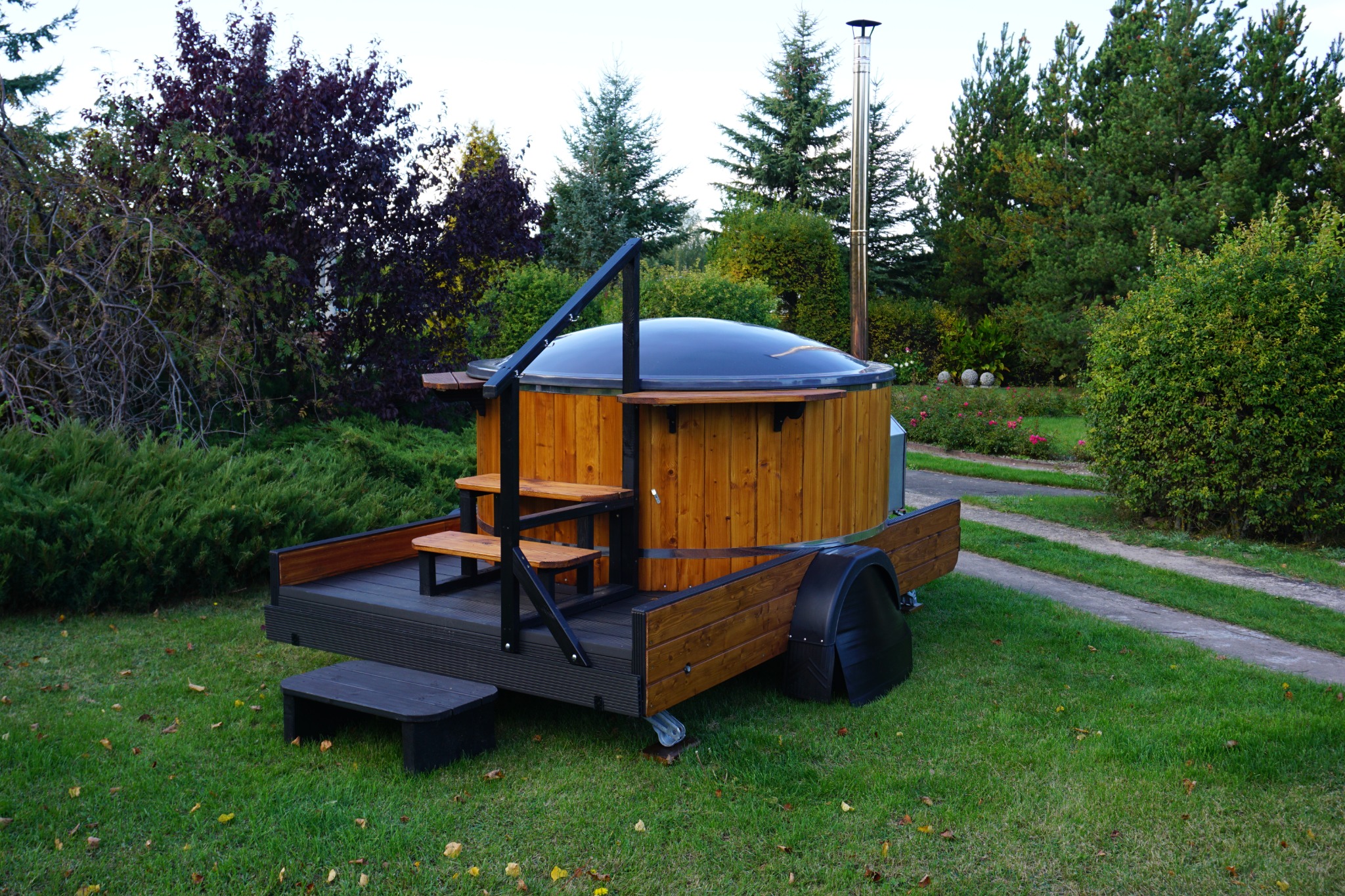 Przenośna, drewniana balia z kominem i schodkami, umieszczona na przyczepie samochodowej, otoczona zielenią.