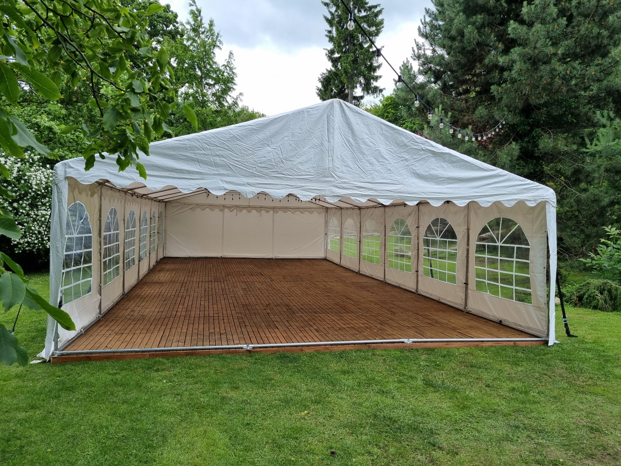 Biały namiot imprezowy z drewnianą podłogą na zielonym trawniku, otoczony drzewami. Widoczne dekoracyjne lampki. Namiot z oknami w kształcie łuków.