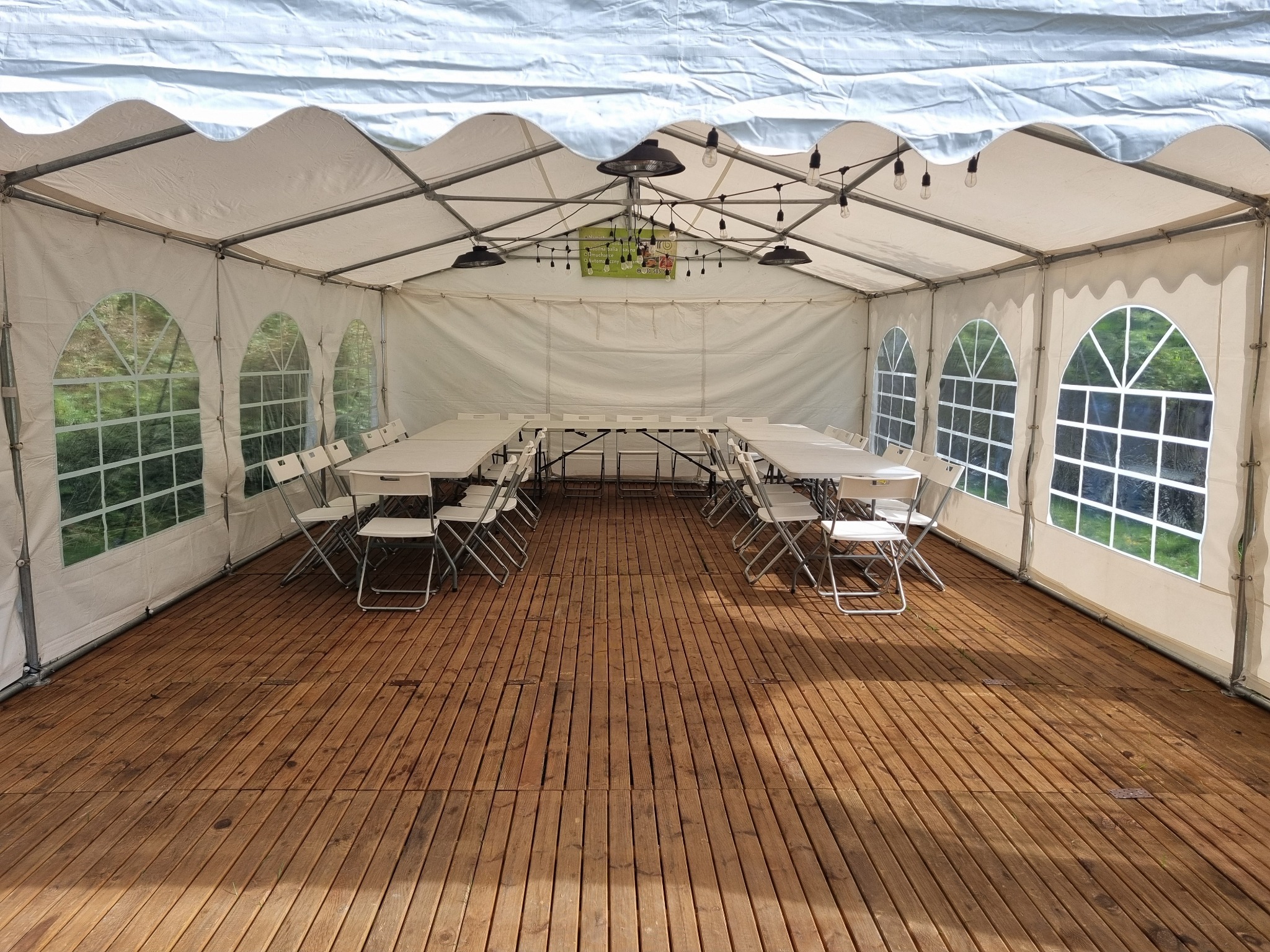 Wnętrze białego namiotu eventowego z drewnianą podłogą, stołami i krzesłami. Oświetlenie w postaci żarówek. Widok na zewnątrz przez okna.