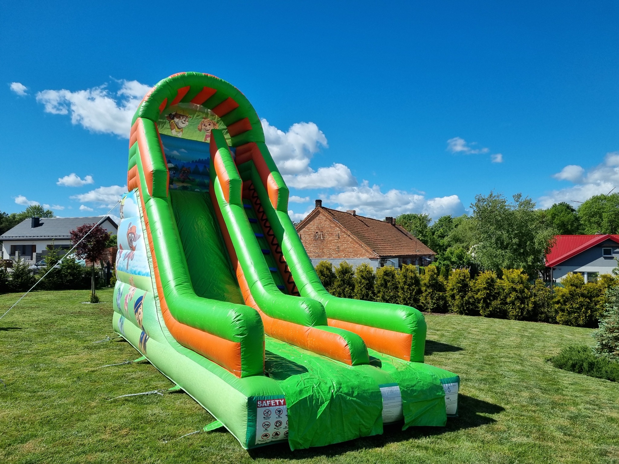 Kolorowa, dmuchana zjeżdżalnia z motywem bajkowym na trawniku, w tle domy i błękitne niebo z chmurami. Widoczne detale mocowania. Bez osób.
