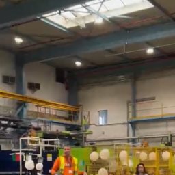 Daria Centka Jendrzej - Event w hali produkcyjnej: mężczyzna w kamizelce odblaskowej przemawia, w tle dekoracje z balonów i osoby w żółtych klatkach. Oświetlenie sufitowe.