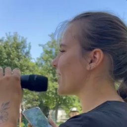 Daria Centka Jendrzej - Kobieta z tatuażem na ramieniu, trzymająca mikrofon i telefon, śpiewa na tle zieleni i błękitnego nieba. Ujęcie z profilu, widoczny fragment sceny.