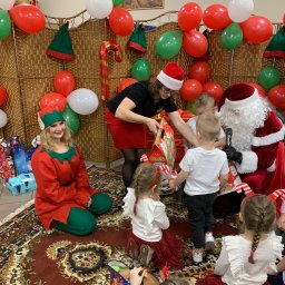 Daria Centka Jendrzej - Święty Mikołaj z elfem rozdają prezenty dzieciom na tle dekoracji z balonów i ozdób świątecznych. Kobieta w czerwonej spódnicy pomaga w rozdawaniu prezentów.