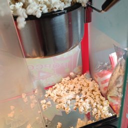 Daria Centka Jendrzej - Automat do popcornu w trakcie pracy, świeżo uprażony popcorn wysypuje się na tacę. Widoczne opakowania na popcorn w tle.