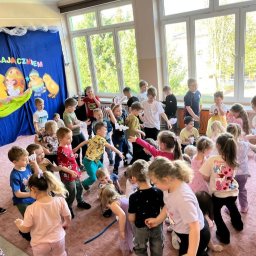 Daria Centka Jendrzej - Radosne dzieci w sali podczas zabawy z okazji Wielkanocy, dekoracje z zajączkami i pisankami w tle, jasne wnętrze, dynamiczne ujęcie.