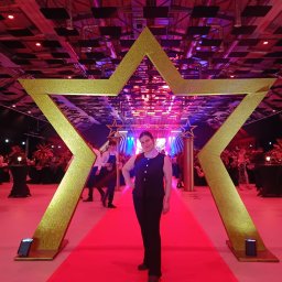 Daria Centka Jendrzej - Kobieta w czarnym stroju pozuje na czerwonym dywanie pod złotą gwiazdą, w tle oświetlona scena i goście. Dekoracja eventowa z efektem czerwonego dywanu.