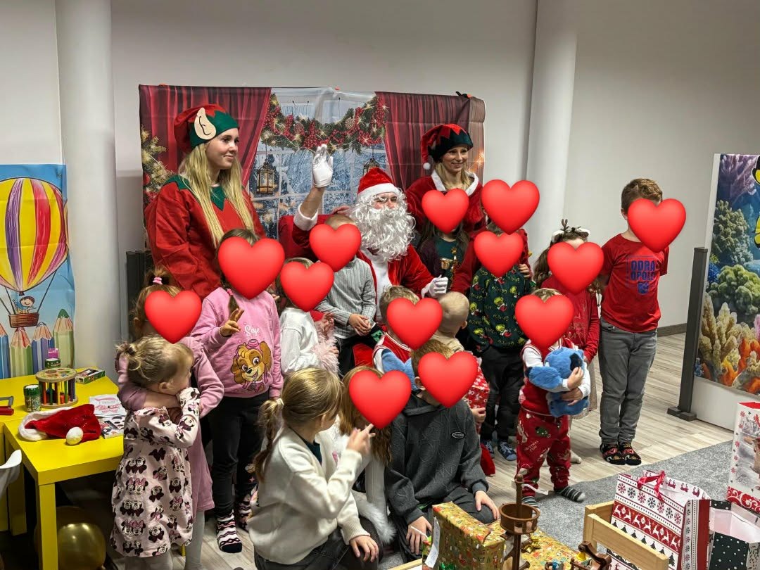 Animatorzy z Wrocławia w strojach elfów i Świętego Mikołaja pozują z grupą dzieci na tle świątecznej dekoracji. Żółty stolik z rekwizytami po lewej.
