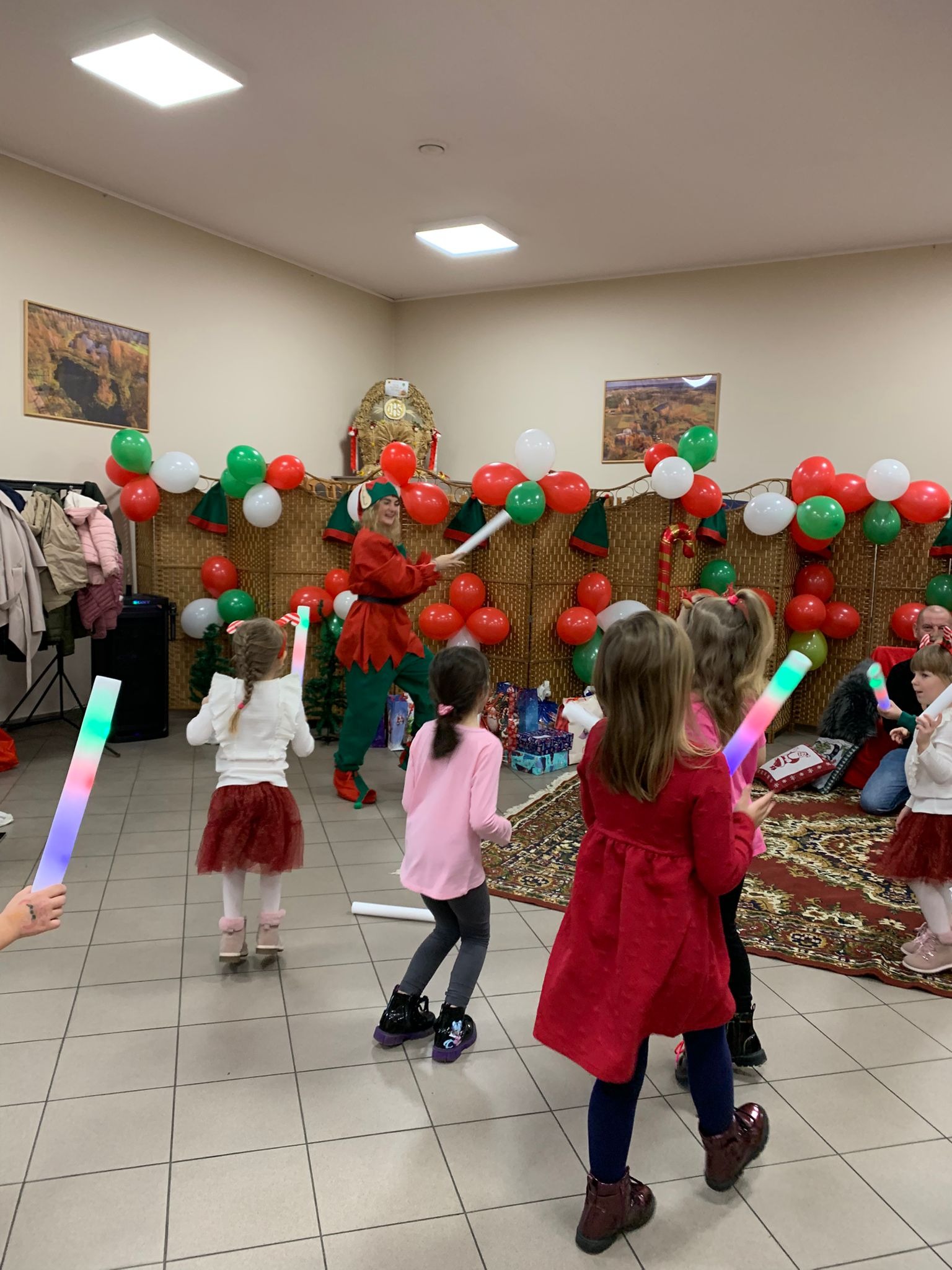 Animator w stroju elfa prowadzi zabawy z dziećmi, trzymając świecące pałeczki. Dekoracje świąteczne z balonów w tle. Ujęcie z perspektywy uczestnika zabawy.