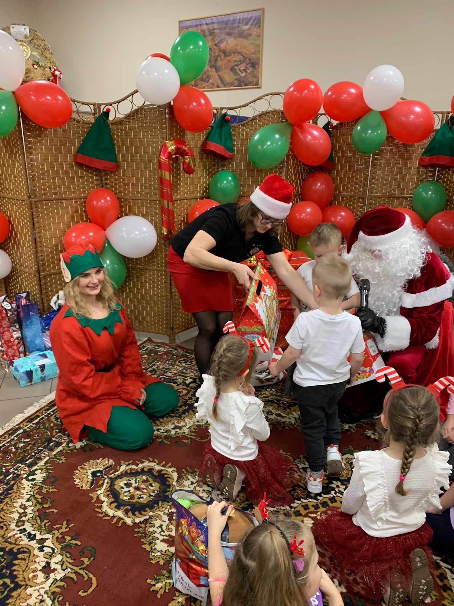 Święty Mikołaj z elfem rozdają prezenty dzieciom na tle dekoracji z balonów i ozdób świątecznych. Kobieta w czerwonej spódnicy pomaga w rozdawaniu prezentów.