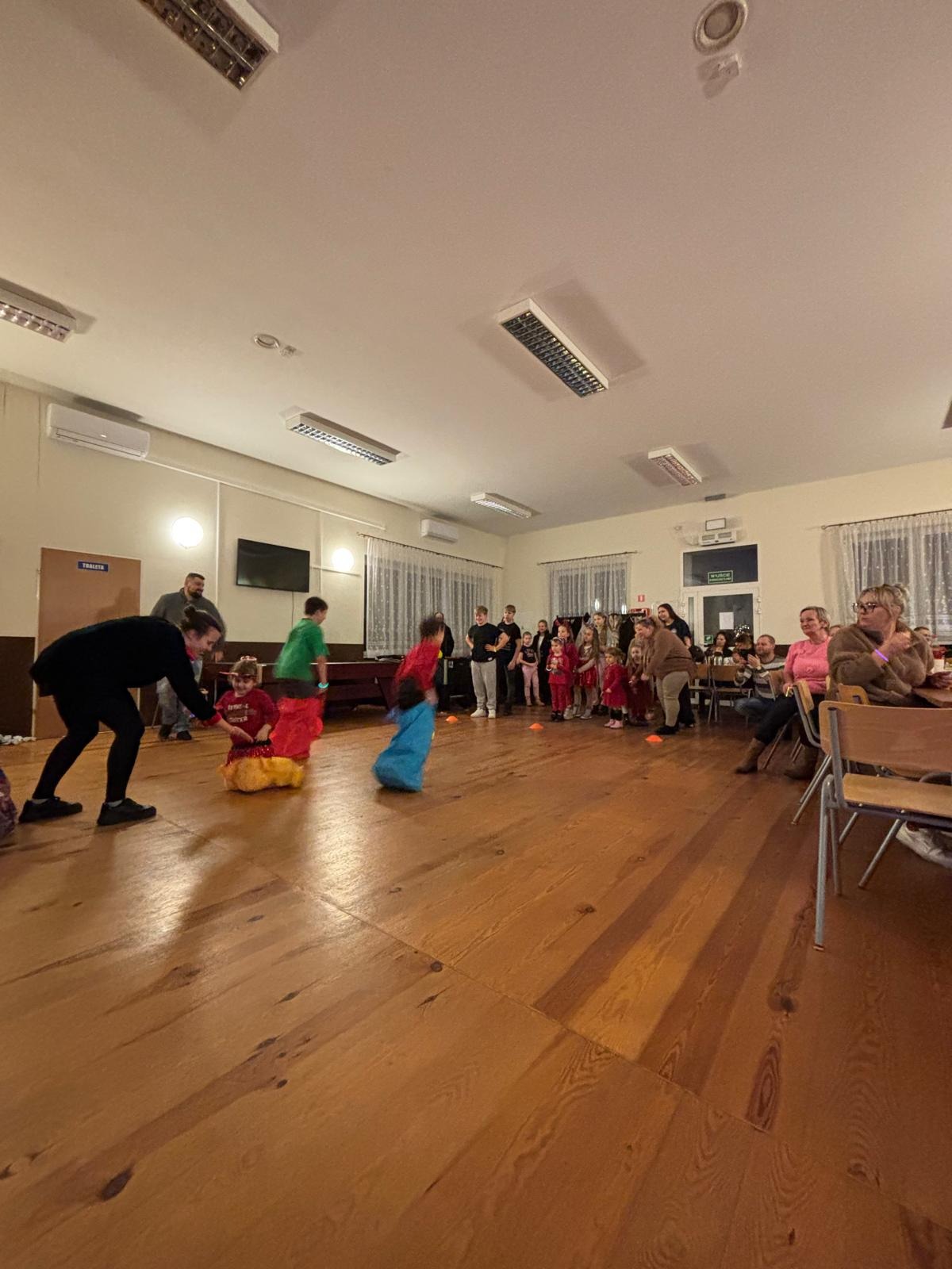 Dzieci w workach skaczą podczas zabawy z animatorem na drewnianej podłodze. W tle grupa osób obserwuje, a w rogu sali siedzą widzowie.