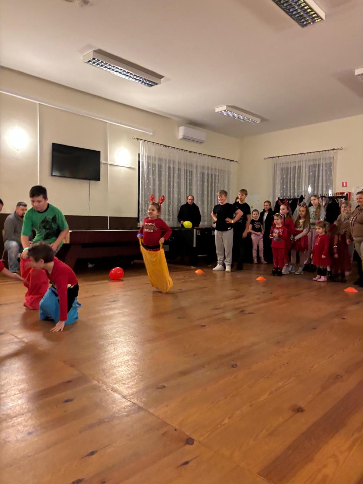 Dzieci biorące udział w zabawie w workach podczas imprezy zorganizowanej w sali. W tle widoczni dorośli i inne dzieci.