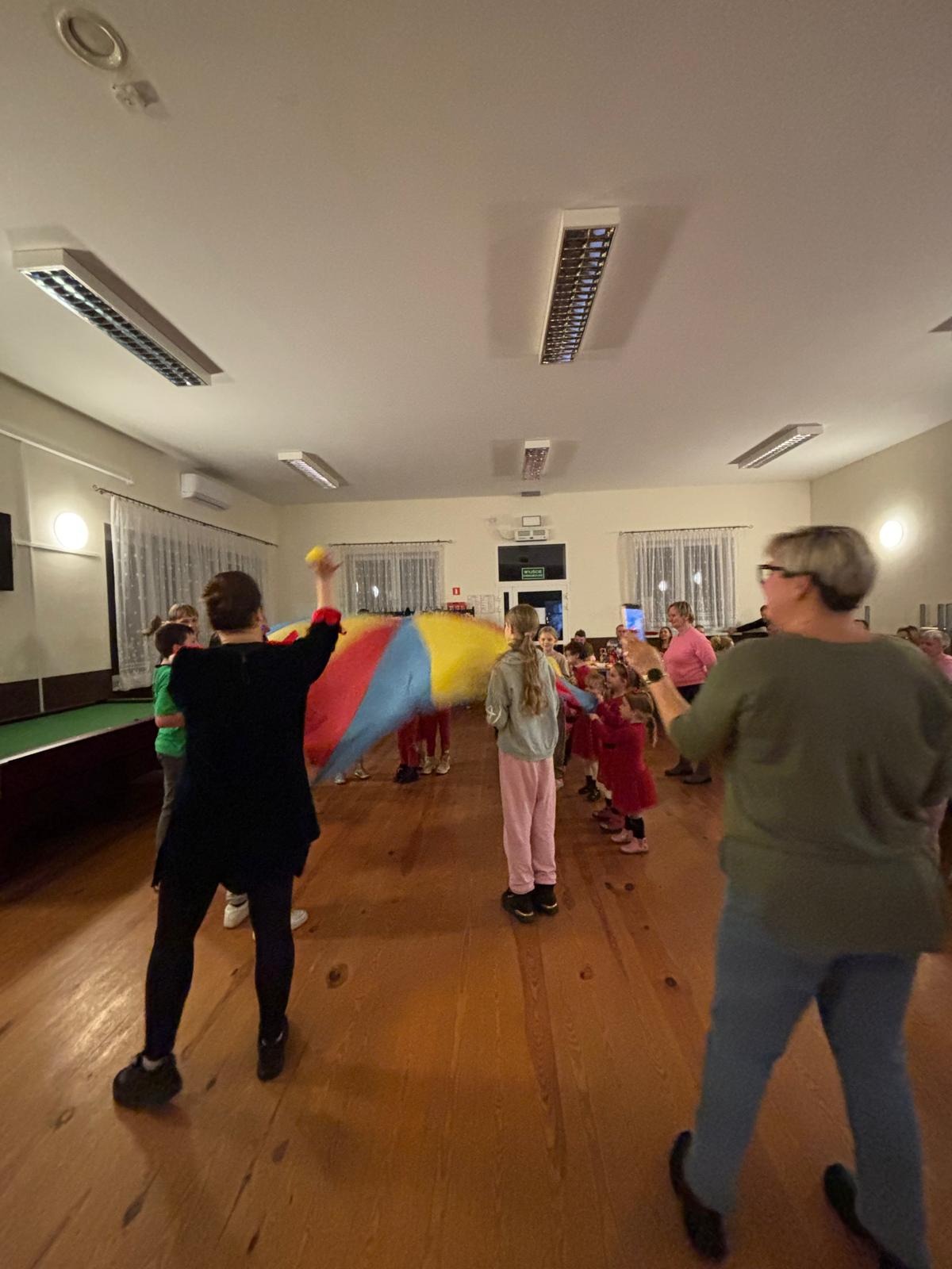 Animator prowadzi zabawy z kolorową chustą animacyjną w przestronnym pomieszczeniu z drewnianą podłogą. Dzieci w kręgu, oświetlenie sufitowe.