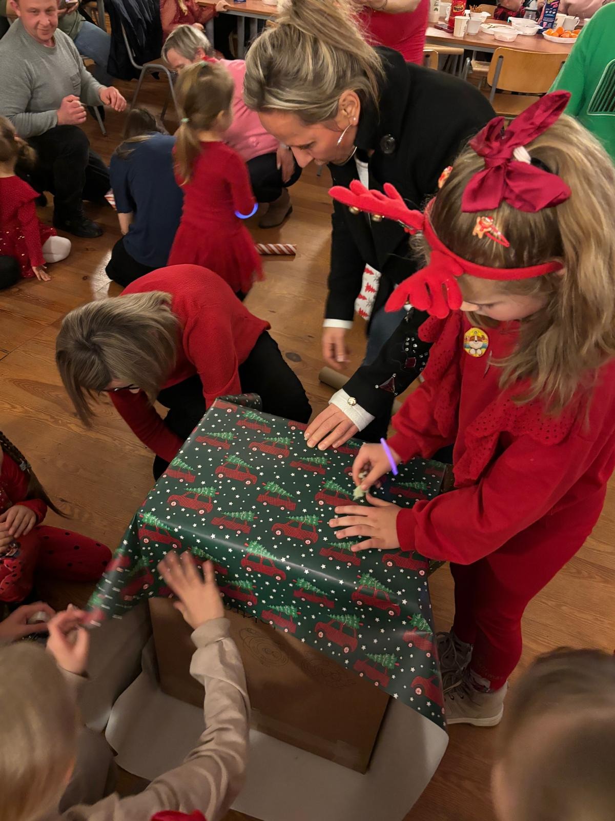 Dzieci w czerwonych strojach, z opaskami reniferów, wspólnie pakują prezent w papier z motywem świątecznych aut na podłodze podczas imprezy.