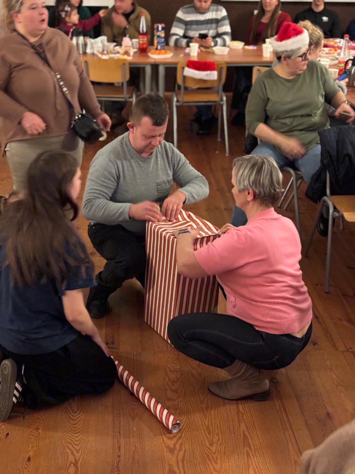 Przygotowania do imprezy we Wrocławiu: grupa osób zawija duży prezent w papier w czerwono-białe paski. W tle widoczny stół z przekąskami i inne osoby.
