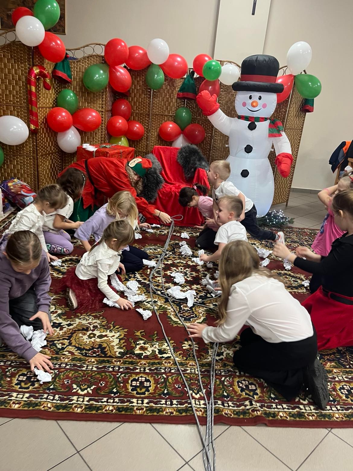 Zabawa dzieci z animatorem w stroju elfa na tle dekoracji świątecznych: dmuchany bałwan, balony i czerwona sofa. Dzieci zbierają papierowe kulki z dywanu.