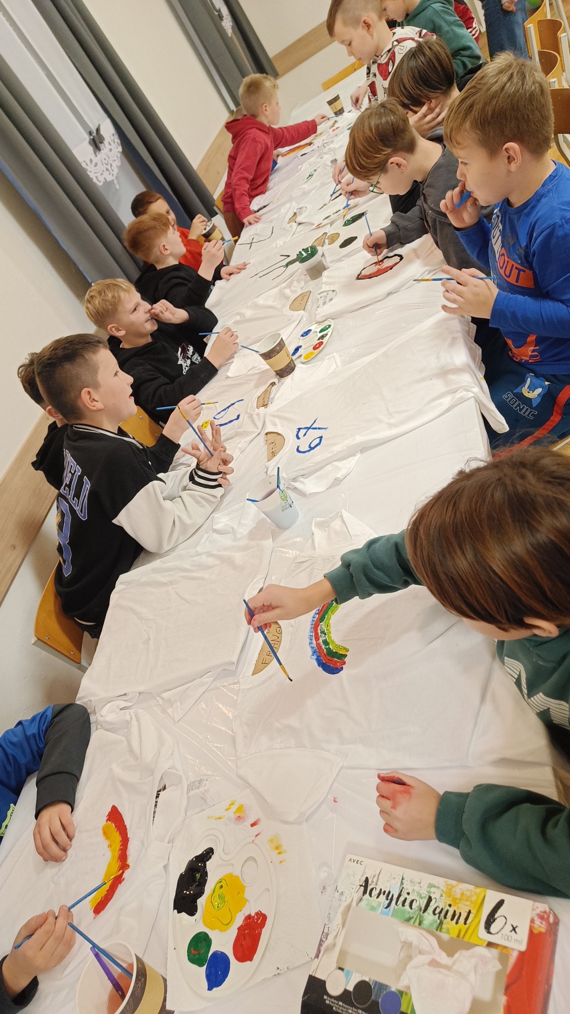 Grupa dzieci maluje farbami na białych koszulkach rozłożonych na długim stole. Widoczne palety z farbami, pędzle i kubki z wodą. Kreatywne zajęcie dla dzieci.