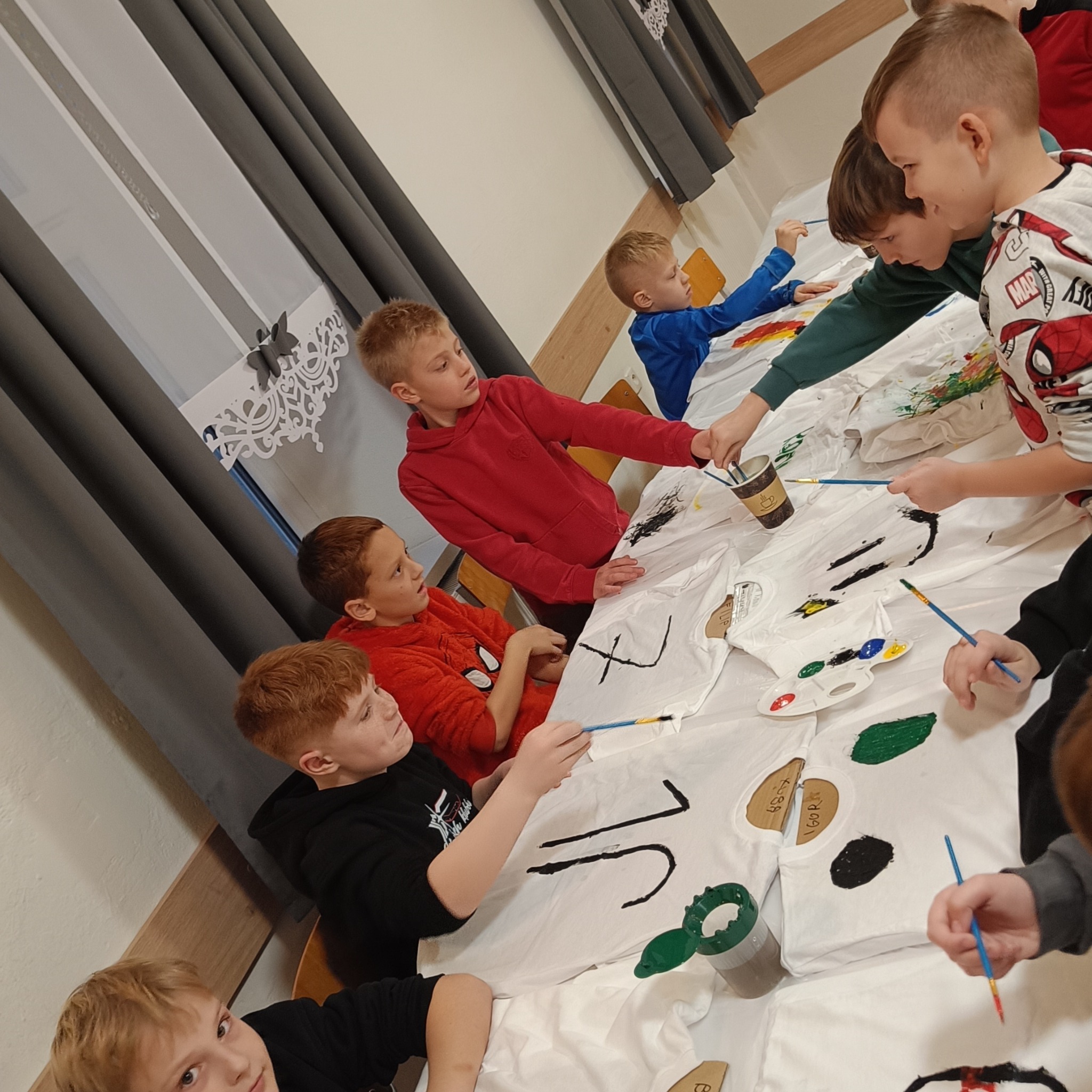 Grupa dzieci maluje farbami na białych koszulkach, siedząc przy stole. Widoczne pędzle, farby i wzory na koszulkach. Kreatywne zajęcia dla dzieci.