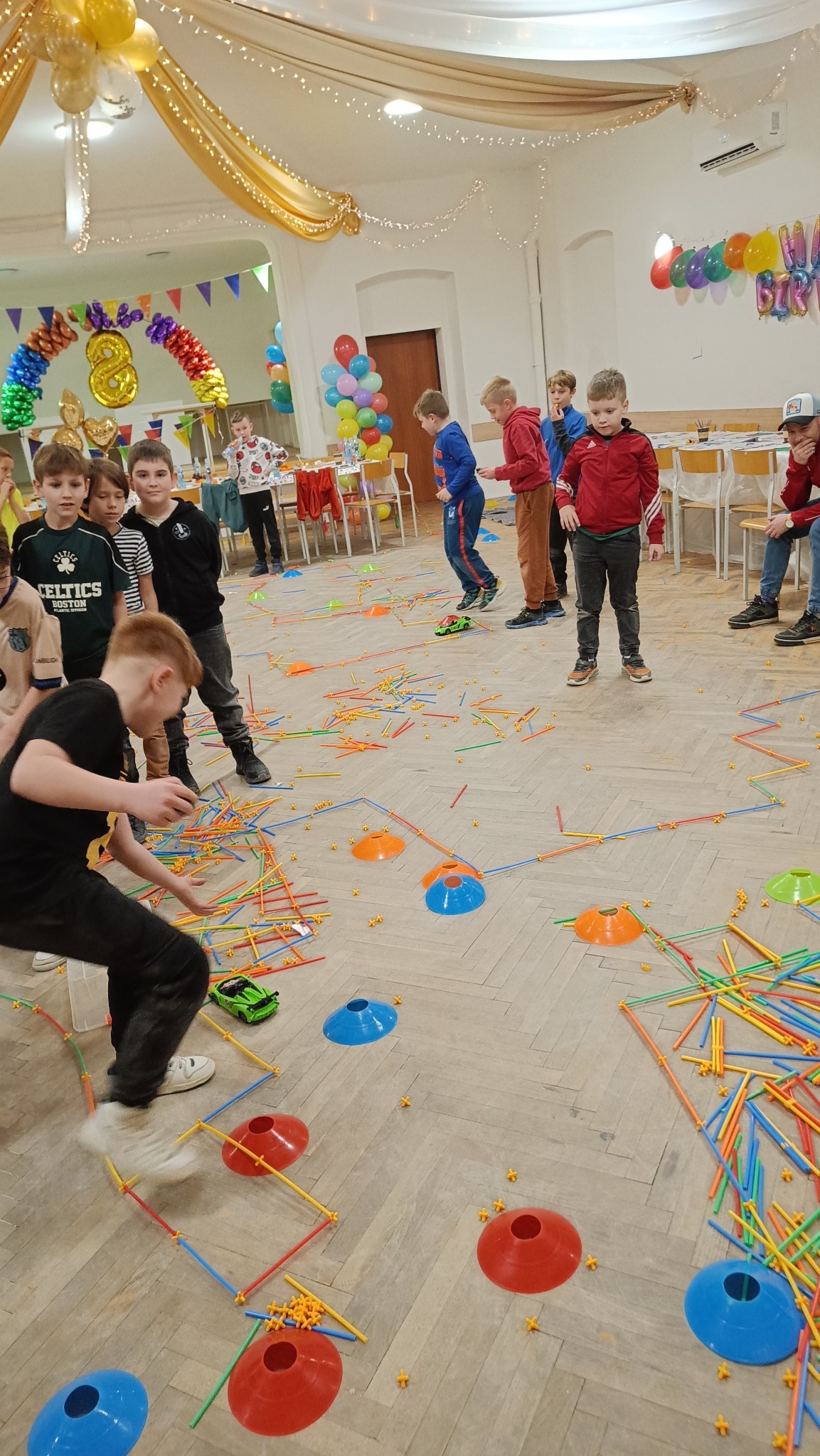 Dzieci bawią się na urodzinach, układając kolorowe konstrukcje z rurek i stożków na parkiecie. W tle dekoracje balonowe i girlandy.
