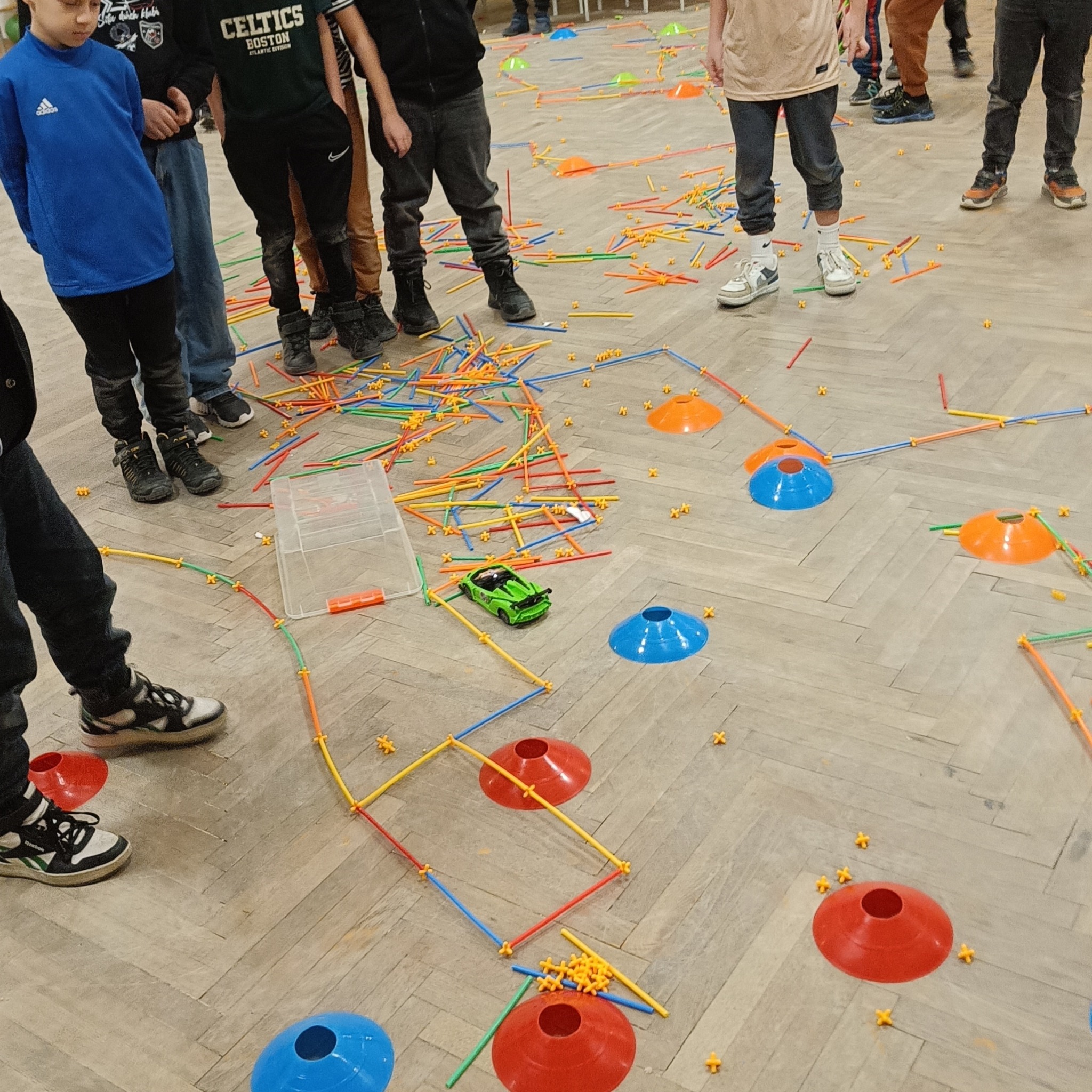 Dzieci budują tor przeszkód z kolorowych rurek i stożków na parkiecie. Na trasie stoi zielony samochodzik. Elementy scenografii imprezy dla dzieci.