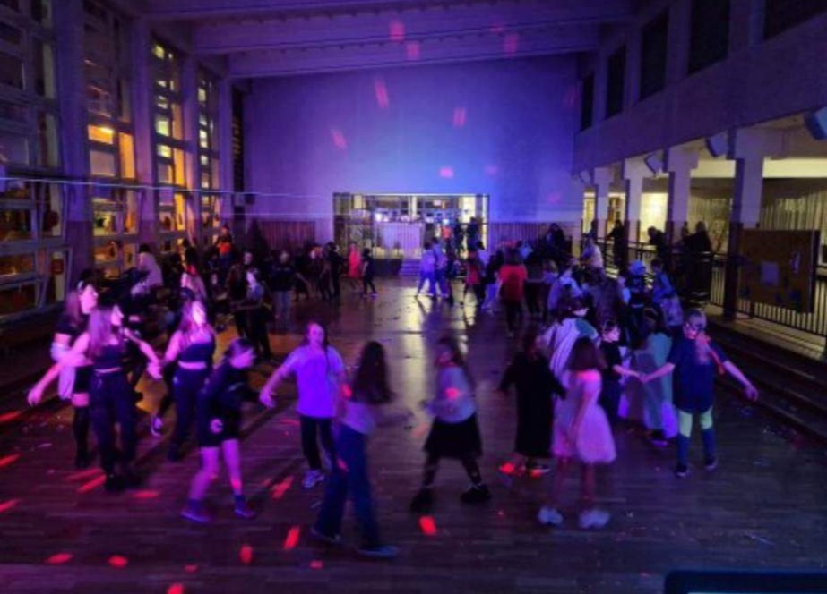 Sala z parkietem wypełniona tańczącymi dziećmi, oświetlona kolorowymi światłami dyskotekowymi. Widoczne okna i balkon na górze. Dynamiczna atmosfera zabawy.