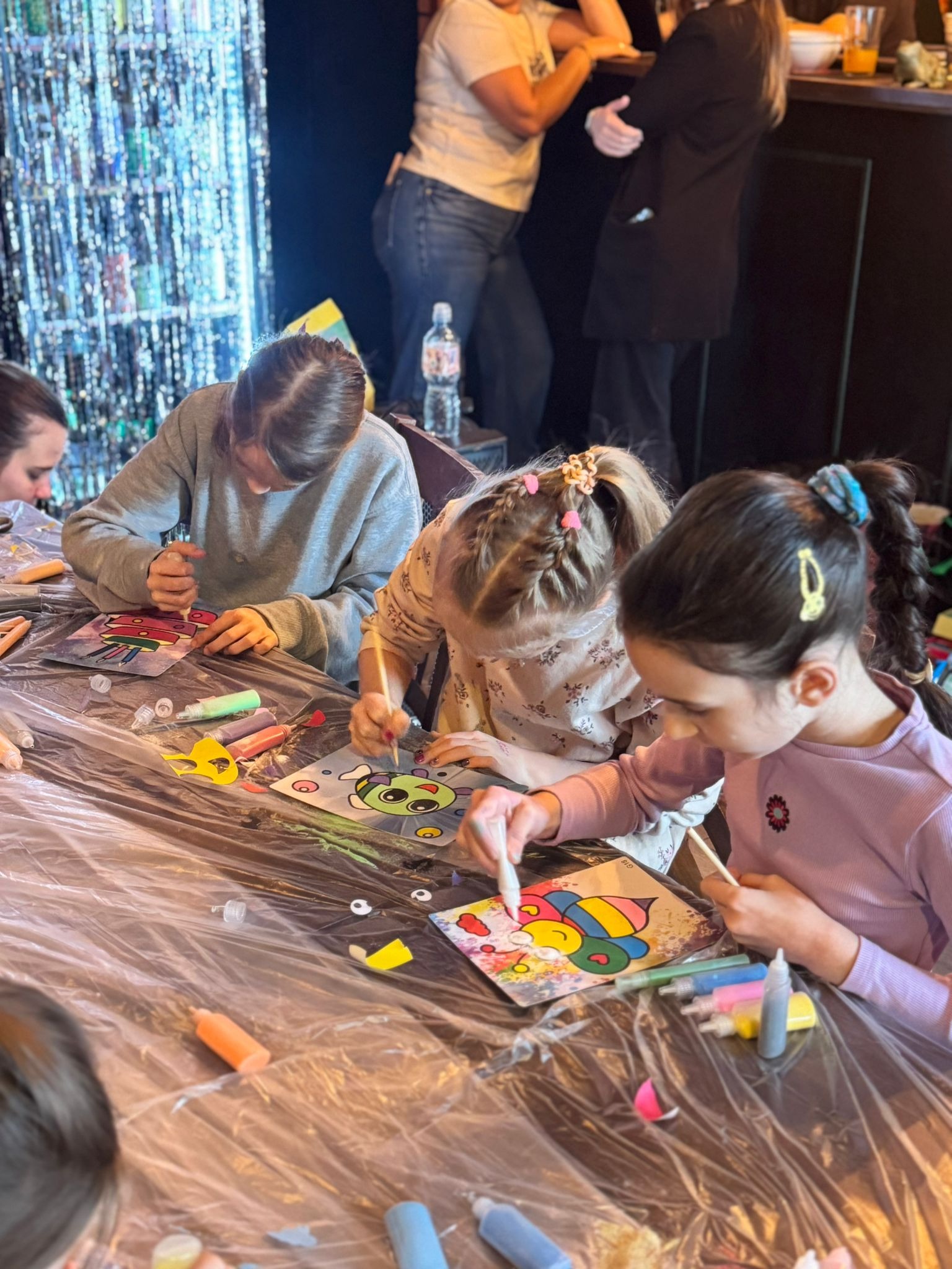 Dzieci malują farbami na szablonach podczas warsztatów artystycznych. Na stole leżą kolorowe farby i pędzle. W tle dekoracja z błyszczących elementów.