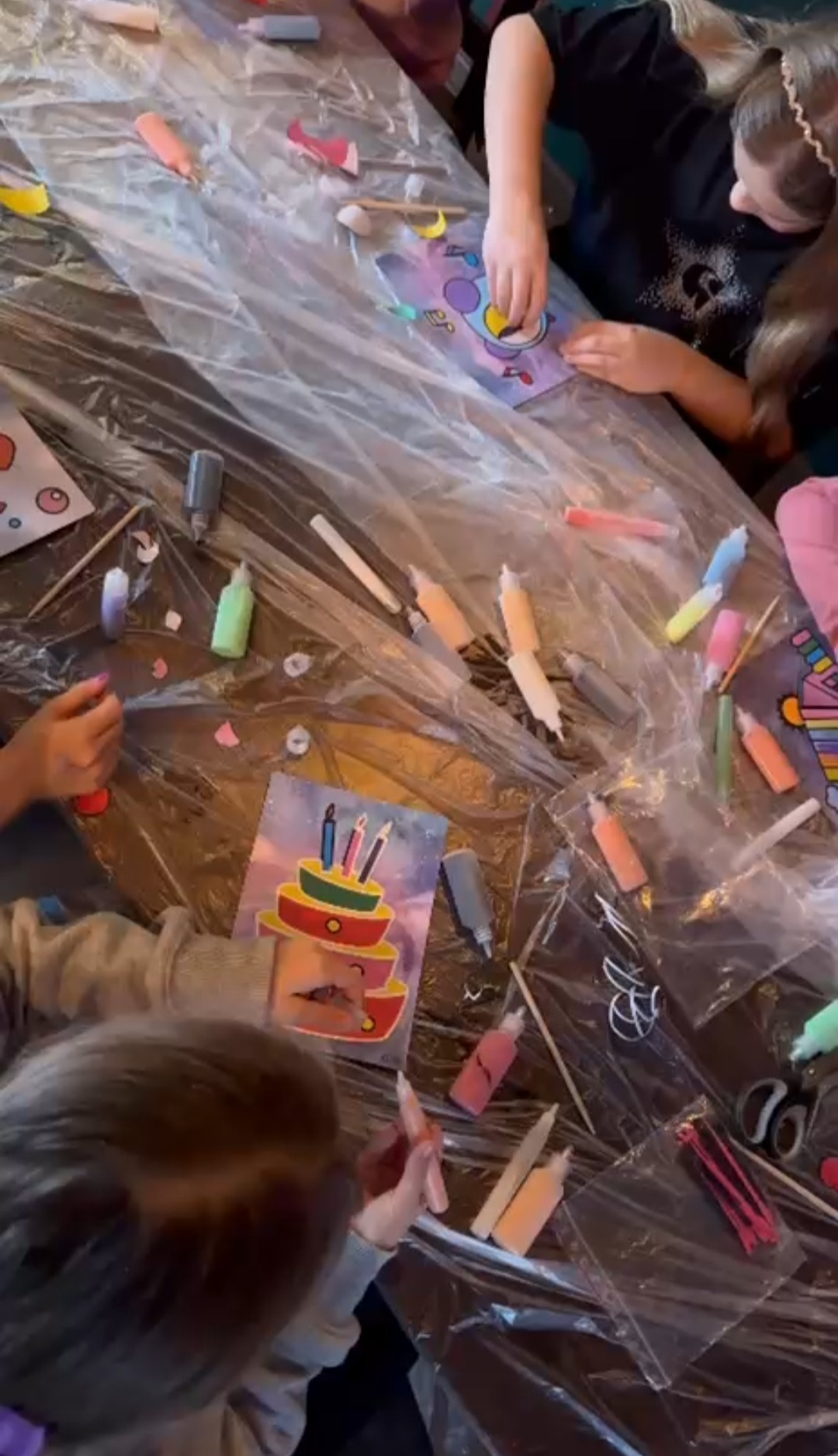 Dzieci dekorują kolorowe kartki z motywami urodzinowymi na stole zabezpieczonym folią, używając farb i pisaków. Kreatywne warsztaty plastyczne.