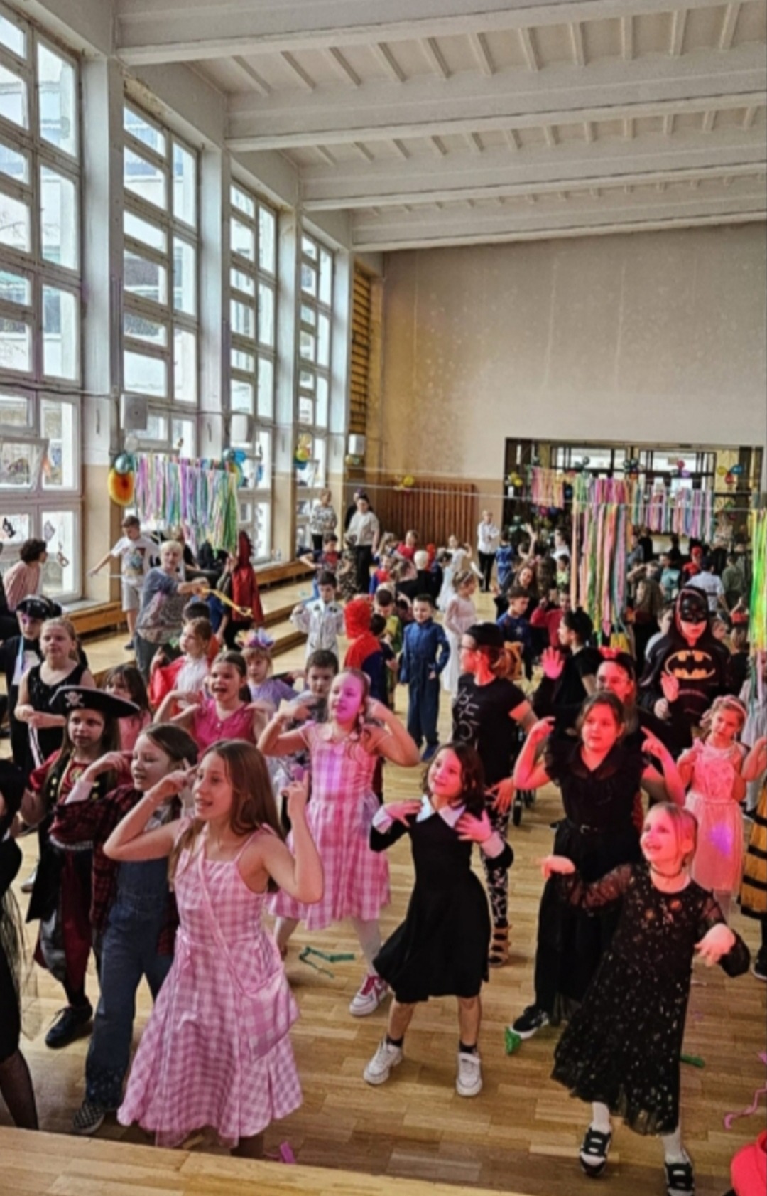 Sala gimnastyczna pełna dzieci w kostiumach karnawałowych, tańczących podczas zabawy. Dekoracje z serpentyn i balonów. W tle duże okna.