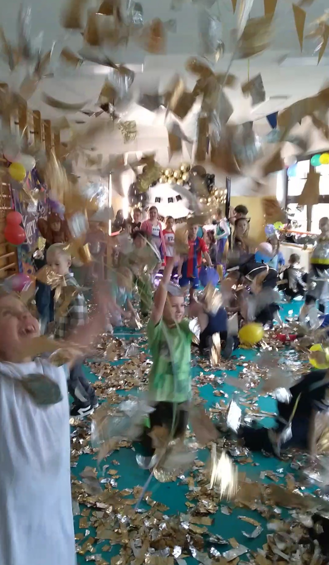 Radosne dzieci w sali udekorowanej balonami i konfetti podczas imprezy urodzinowej. Chłopiec w zielonej koszulce podnosi ręce w geście radości.