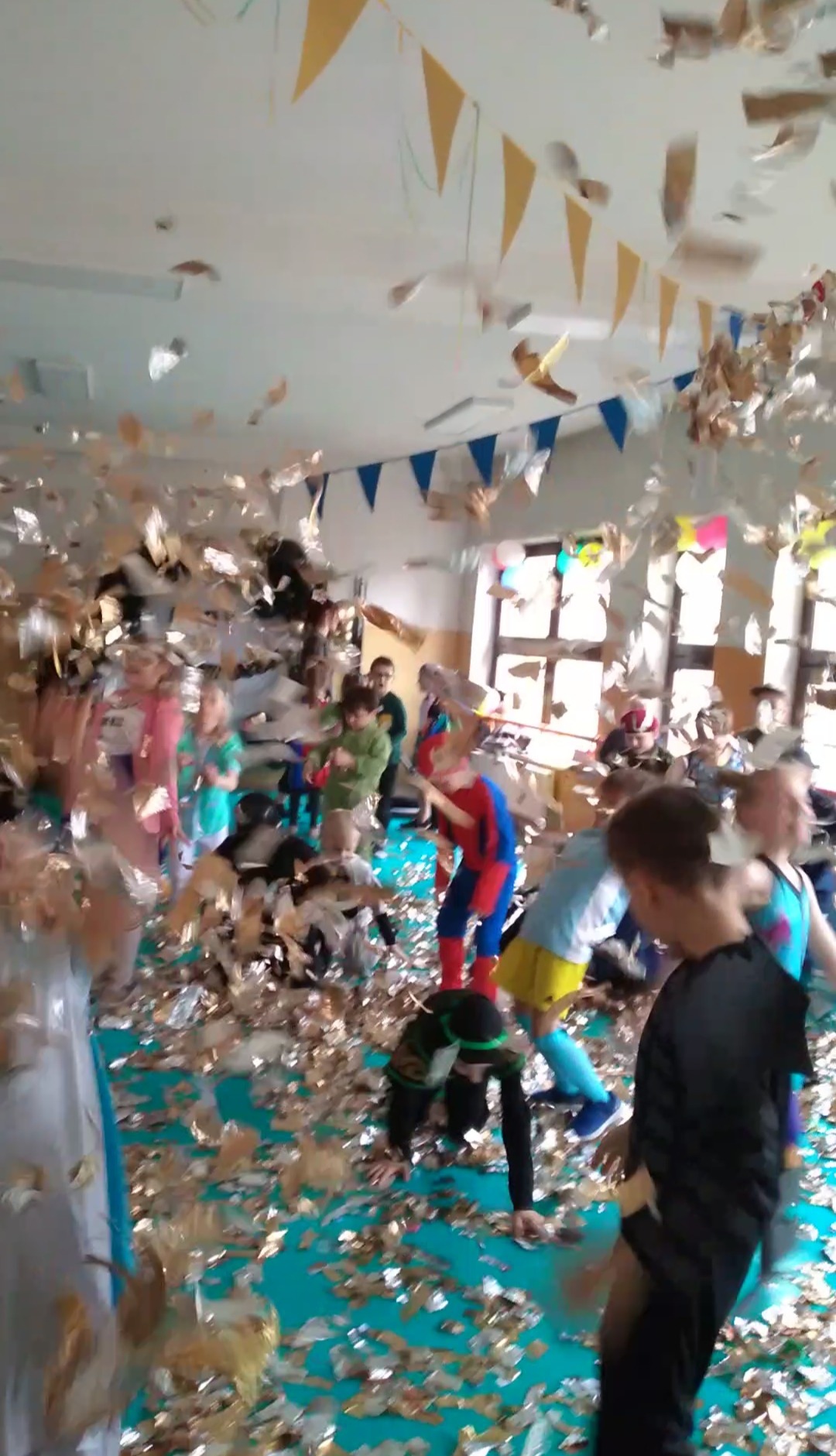 Sala udekorowana girlandami, wypełniona bawiącymi się dziećmi w kostiumach i konfetti, tworząc radosną atmosferę imprezy.