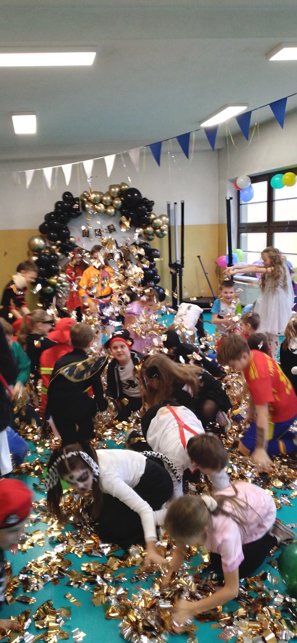 Sala gimnastyczna udekorowana balonami i girlandami, dzieci w kostiumach bawią się w konfetti podczas imprezy z animatorem.