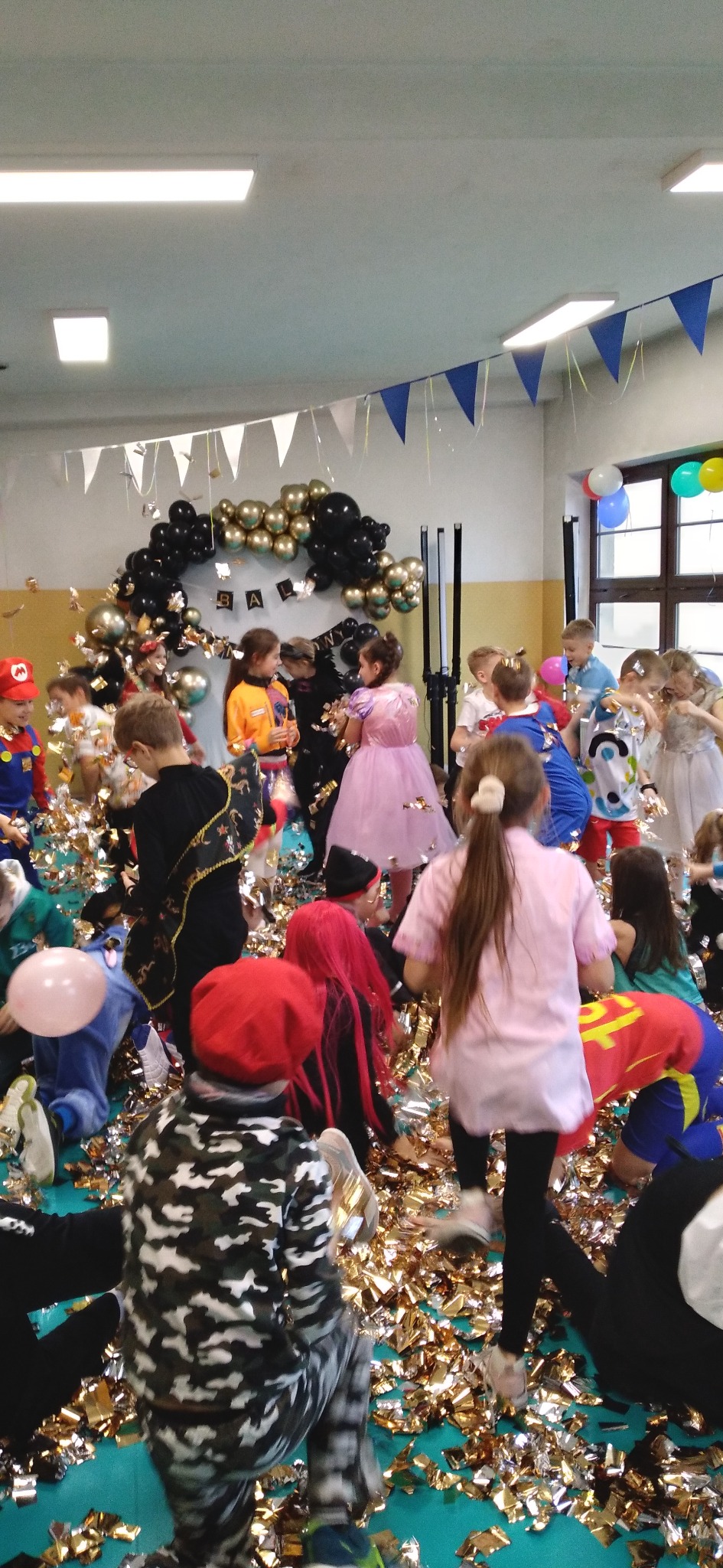 Sala balowa wypełniona konfetti, dzieci w kostiumach bawią się pod dekoracją z balonów. Ujęcie z perspektywy uczestnika zabawy, widoczne detale strojów i radość.