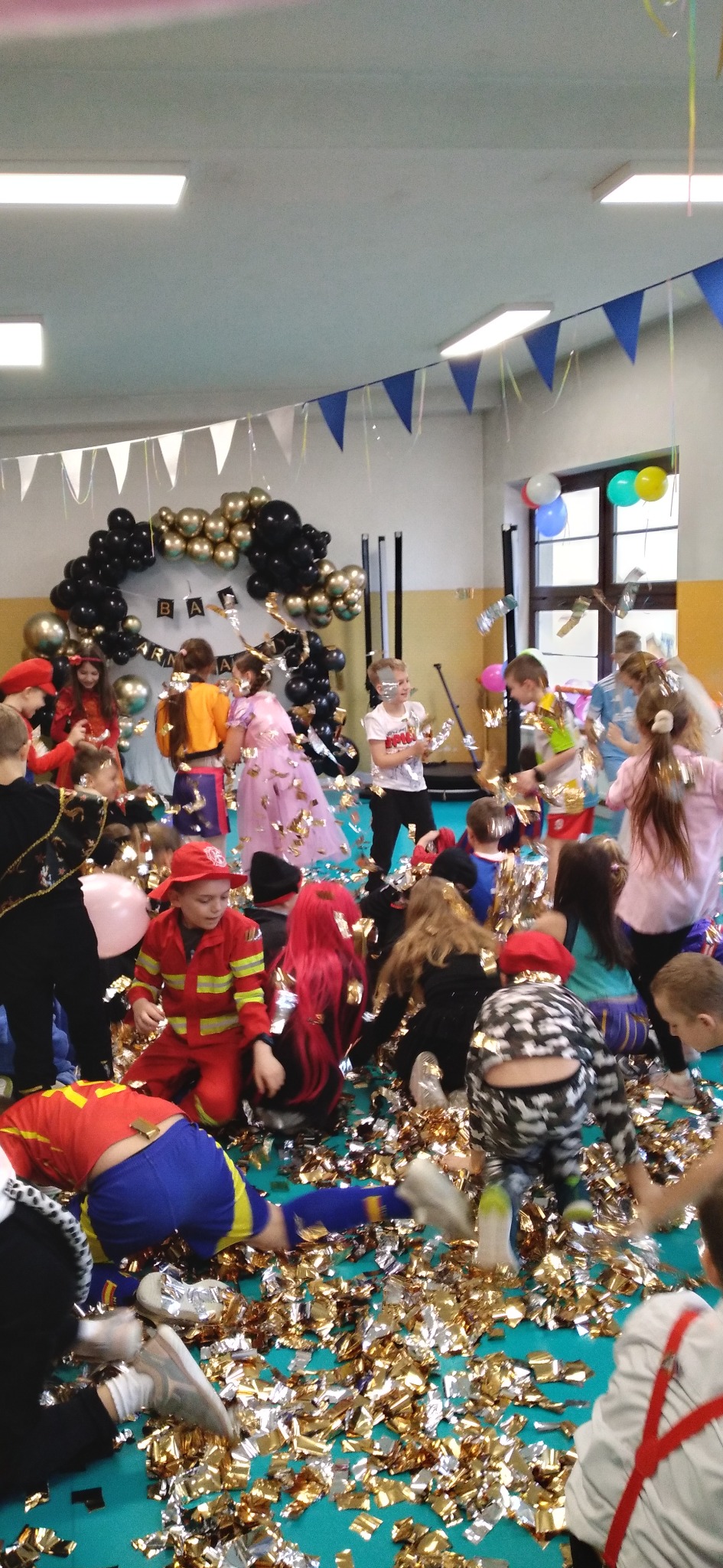 Urodziny dziecka w sali gimnastycznej: dzieci w kostiumach bawią się w konfetti, w tle dekoracja z balonów i girlandy.