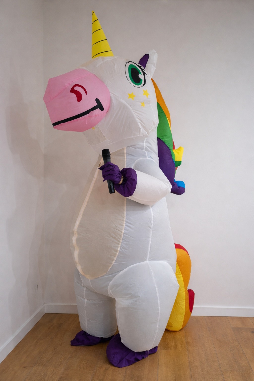 Nadmuchiwany kostium jednorożca z tęczową grzywą, trzymający mikrofon, stojący w rogu pokoju z drewnianą podłogą i białymi ścianami. Kostium ma żółty róg i różowy pysk.