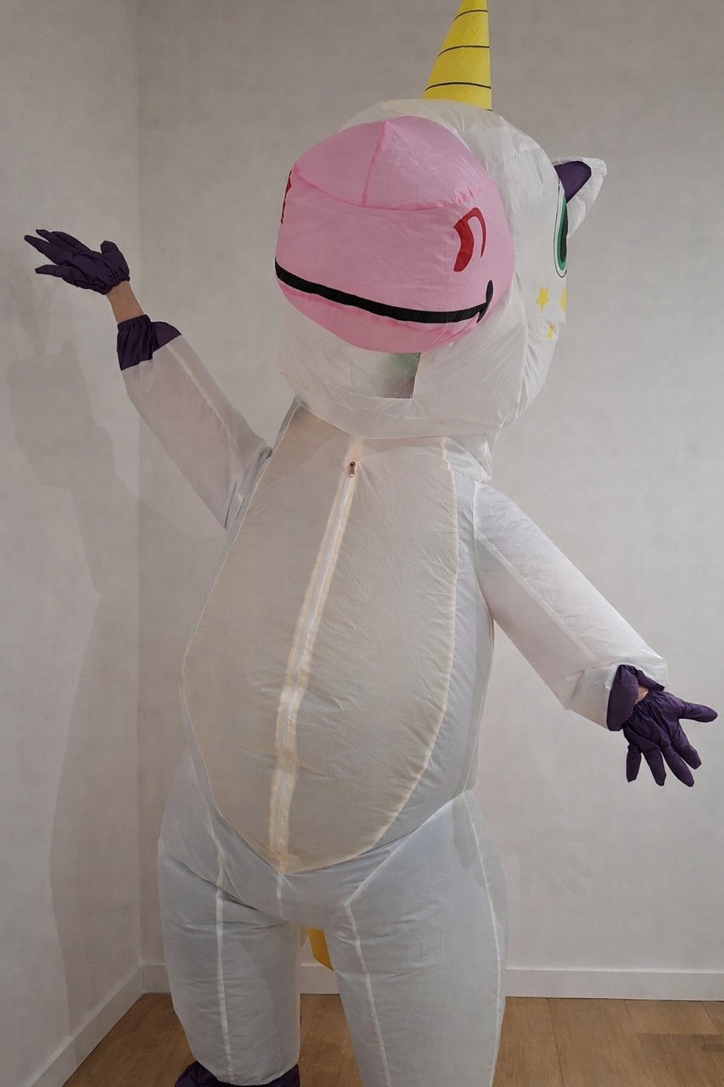 Osoba w dmuchanym kostiumie jednorożca z różową głową i żółtym rogiem, pozująca na tle jasnej ściany. Widoczne fioletowe rękawiczki i zamek błyskawiczny na kostiumie.