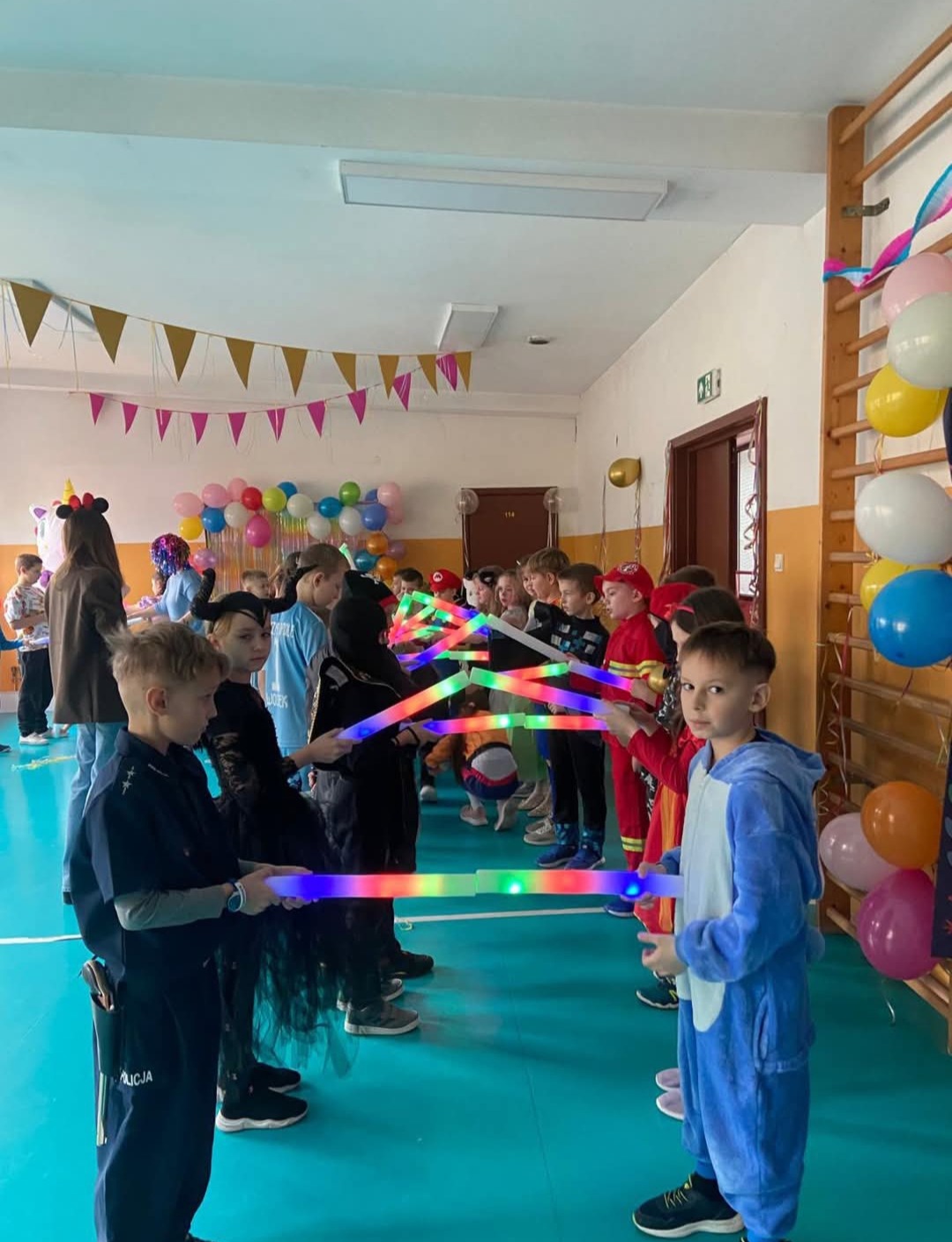 Dzieci w kostiumach, trzymające świetlne pałeczki, tworzą szpaler na tle sali udekorowanej balonami i girlandami. Uczestnicy zabawy w różnorodnych przebraniach.
