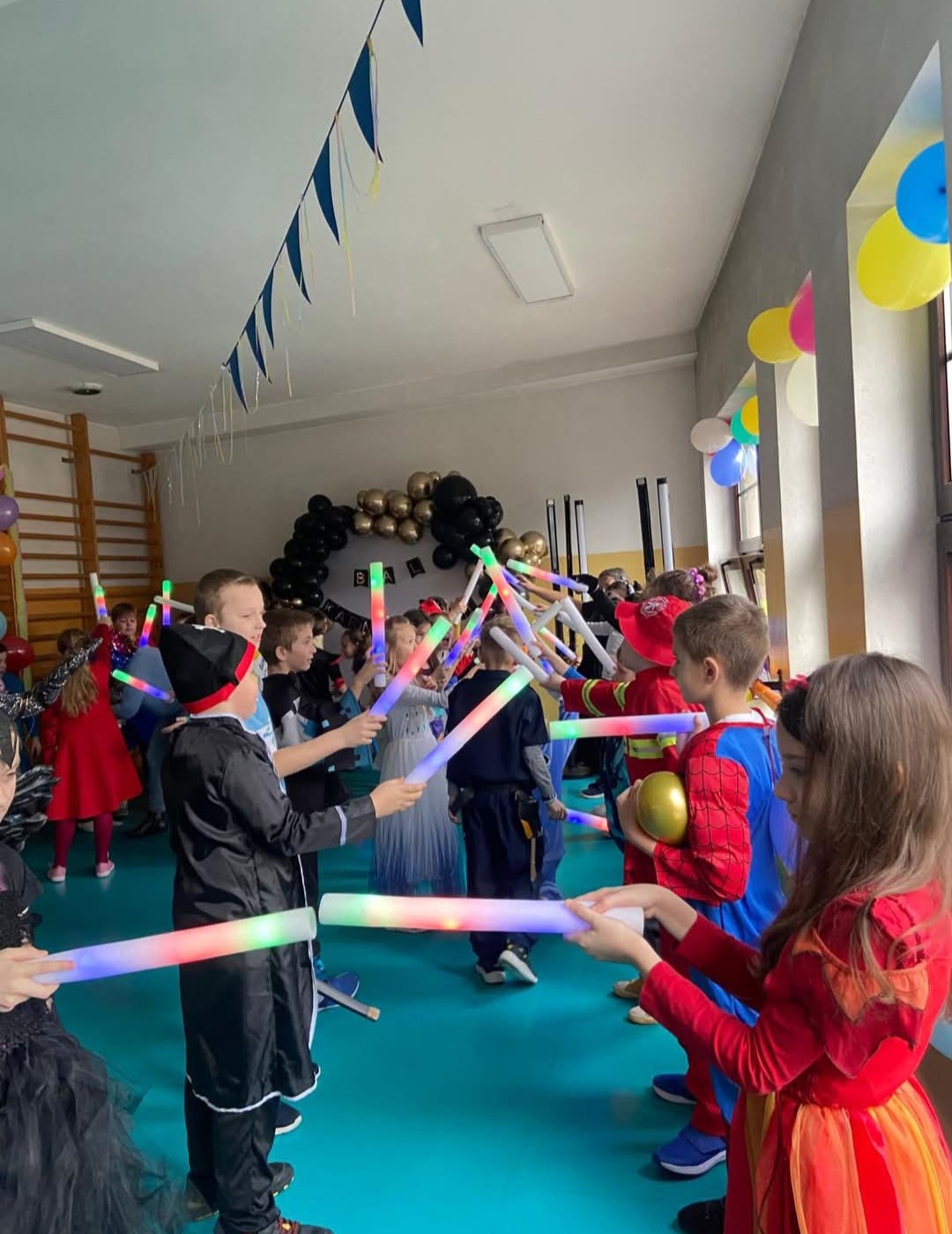 Dzieci w kostiumach bawią się świecącymi pałkami na tle dekoracji balonowych i girland, podczas imprezy zorganizowanej w sali gimnastycznej.