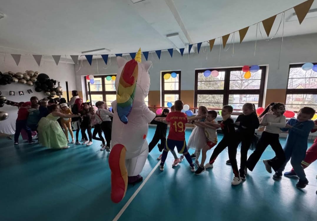 Animator w stroju jednorożca prowadzi zabawę przeciągania liny z grupą dzieci na tle sali gimnastycznej udekorowanej balonami i girlandami.