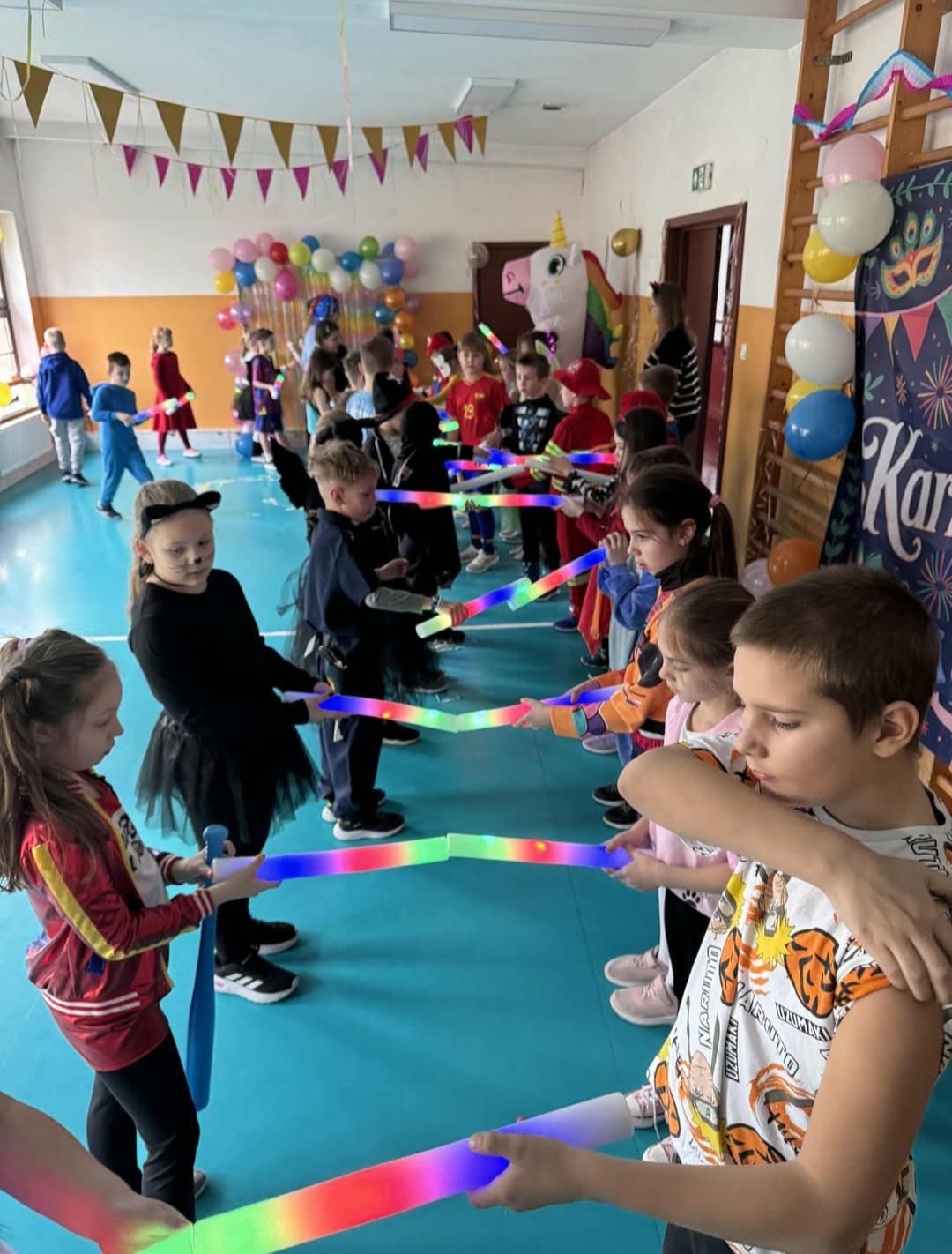 Karnawałowy bal przebierańców w sali gimnastycznej: dzieci w kostiumach z kolorowymi, świecącymi pałkami tworzą szpaler. Dekoracje balonowe i jednorożec w tle.