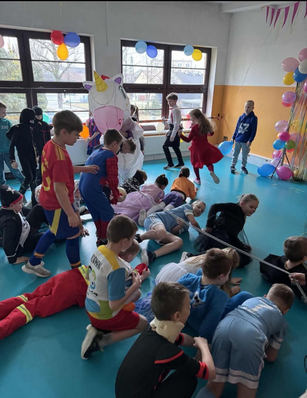 Dzieci w kostiumach bawią się na podłodze w sali udekorowanej balonami, w tle okna i osoba w kostiumie jednorożca. Impreza karnawałowa dla dzieci.