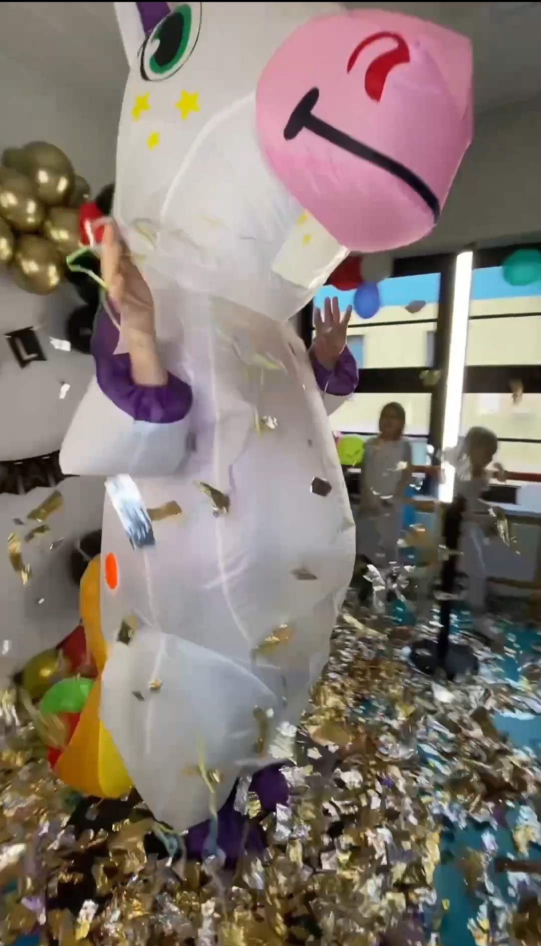 Animator w dmuchanym kostiumie jednorożca, unoszący ręce w geście powitania, otoczony złotym konfetti podczas imprezy dla dzieci. W tle dzieci i balony.