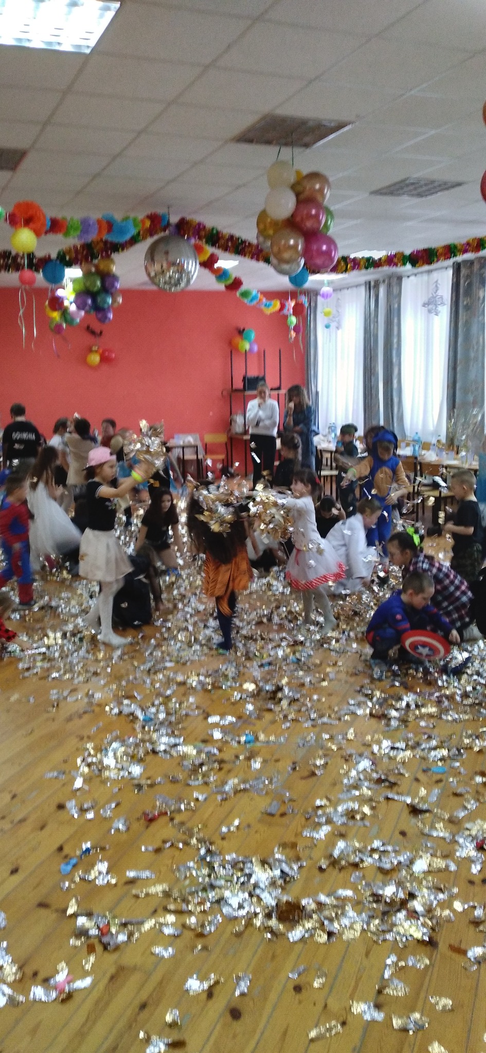 Sala udekorowana balonami i konfetti, dzieci w kostiumach bawią się podczas imprezy. Widoczna kula dyskotekowa i dekoracje.