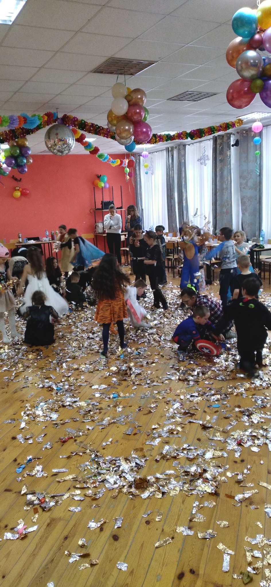 Sala po imprezie dla dzieci, parkiet zasypany konfetti, w tle dzieci w kostiumach i dekoracje z balonów.
