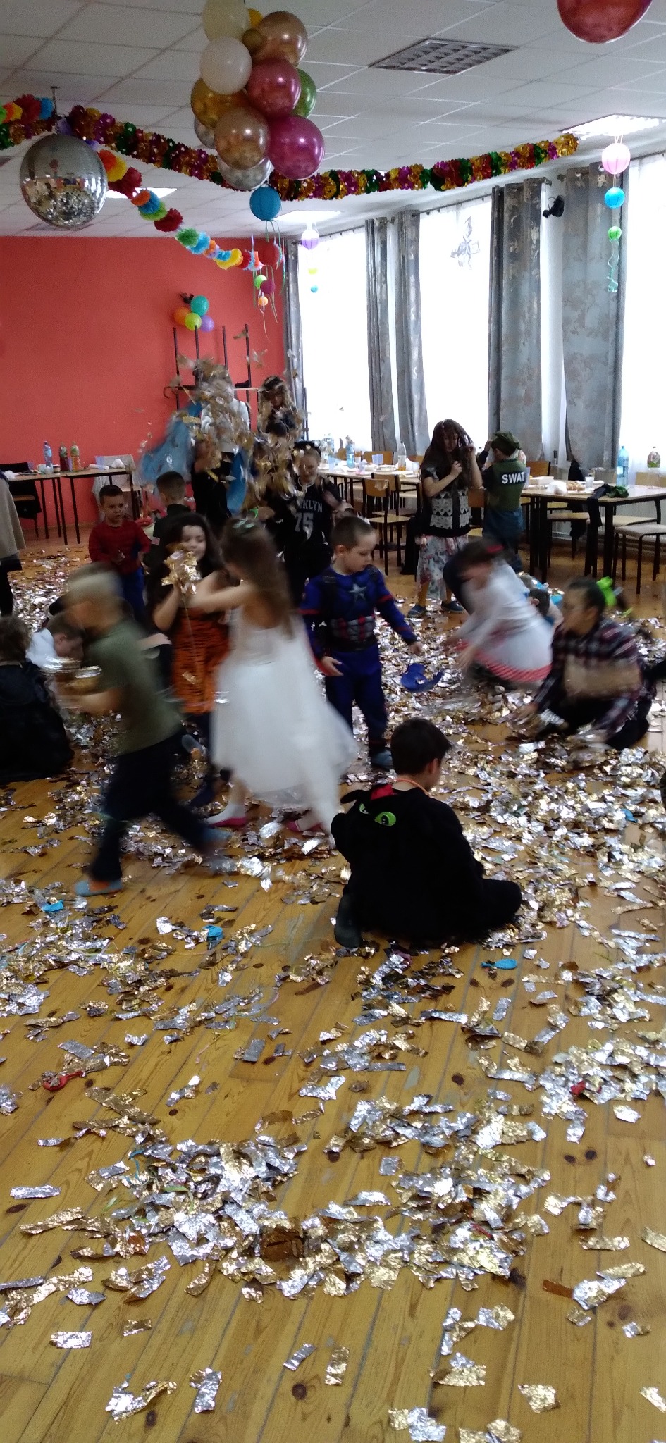 Dzieci bawią się na parkiecie zasypanym konfetti podczas imprezy. Dekoracje z balonów i girlandy. Uczestnicy w kostiumach.