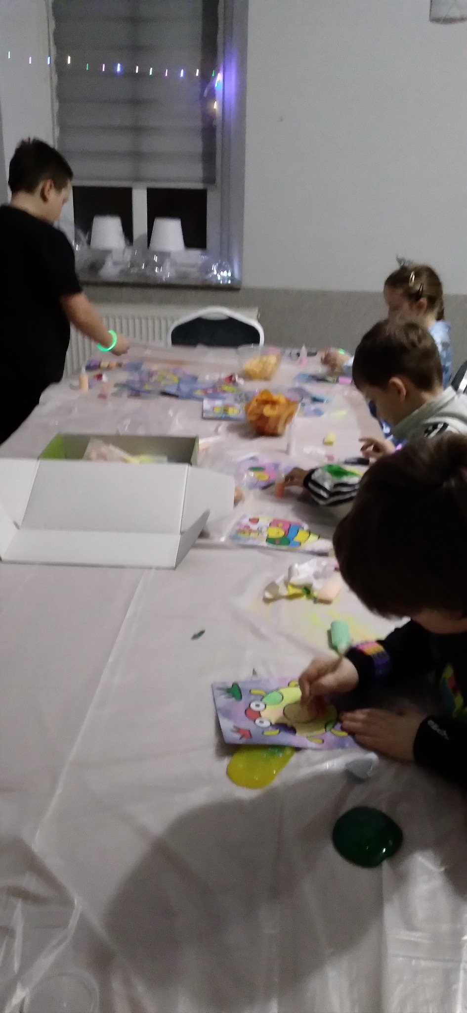 Dzieci przy stole, malują kolorowanki i bawią się slime podczas animacji urodzinowej. Widoczne pudełka, dekoracje i oświetlenie.