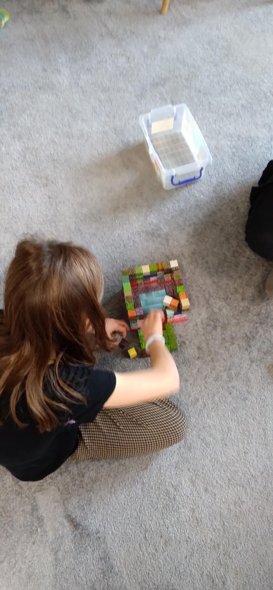 Dziewczynka siedzi na podłodze i buduje konstrukcję z kolorowych klocków. Obok pojemnik na klocki. Ujęcie z góry, widoczne włosy i ręce dziecka.