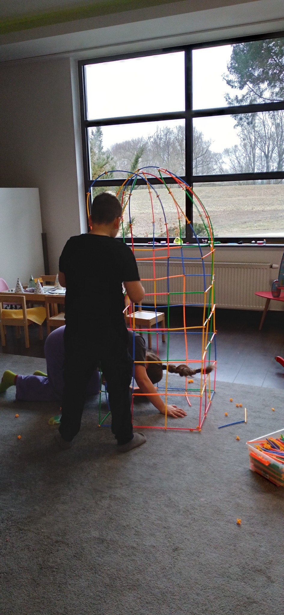 Dzieci budują kolorową konstrukcję z rurek w pokoju z dużym oknem. Widoczne elementy konstrukcyjne i pudełko z klockami. Zabawa w pomieszczeniu.