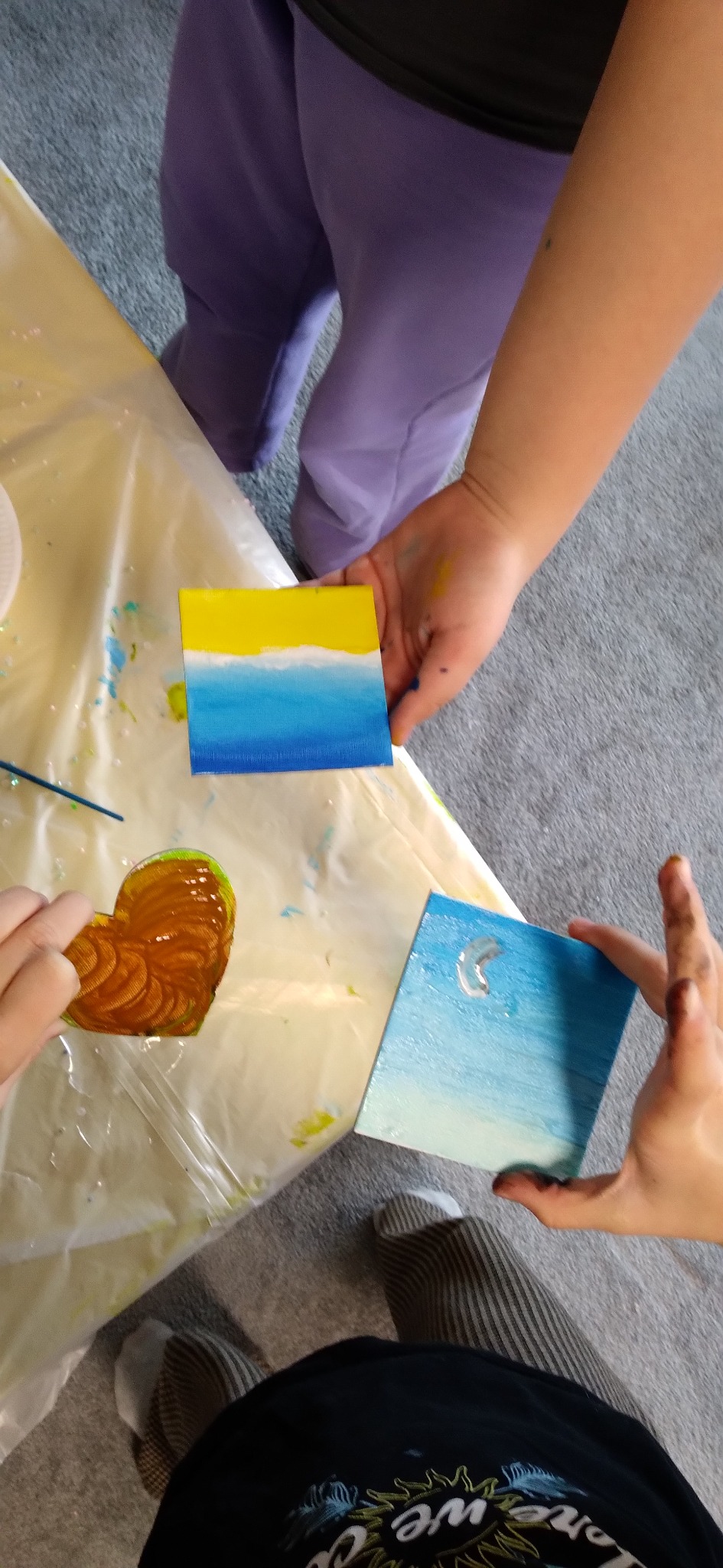 Dzieci prezentują własnoręcznie malowane, małe obrazy na płótnie: pejzaż morski, słońce nad wodą i serce. Kreatywne warsztaty plastyczne dla dzieci.