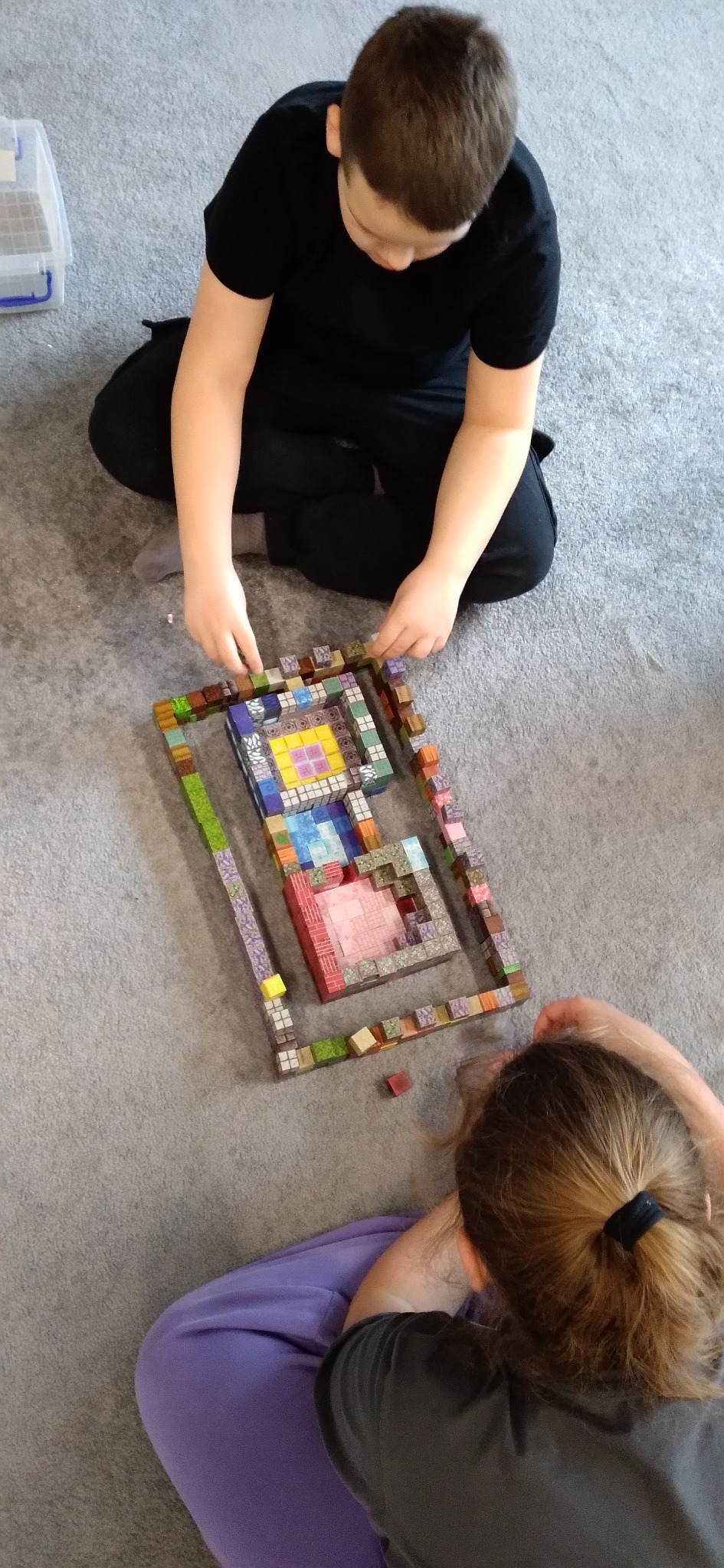 Dzieci bawią się klockami, tworząc konstrukcję przypominającą budowlę z gry Minecraft. Ujęcie z góry, widoczne ręce układające klocki i fragmenty ubrań dzieci.